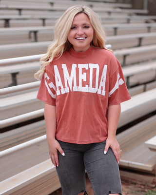 A smiling blonde woman stands in front of outdoor bleachers wearing a terracotta short-sleeve graphic T-shirt with 'GAMEDAY' printed in white across the chest, paired with dark gray ripped jeans.