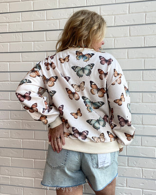 A woman viewed from behind wearing a cream-colored crewneck sweatshirt covered in a colorful butterfly print, paired with light-wash denim shorts, standing against a white brick wall.