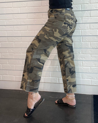 A person wearing camouflage cargo pants, a black tank top, and black slide sandals, posing against a white brick wall.