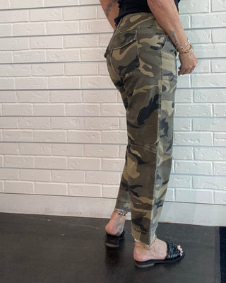A woman wearing camouflage cargo pants and a black tank top, shown in a side-profile mirror selfie against a white brick wall.