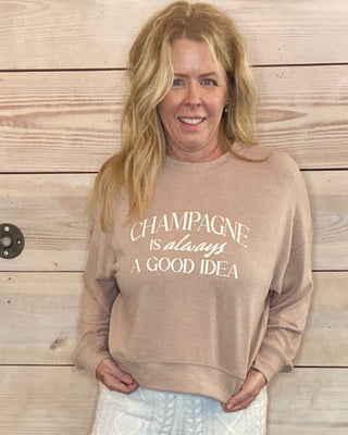 Woman wearing a beige sweatshirt with text and white pants against a wooden wall.