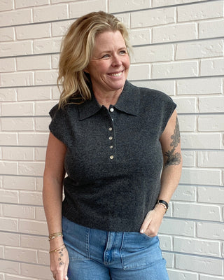 Woman wearing a dark sleeveless sweater and blue jeans standing against a light brick wall.