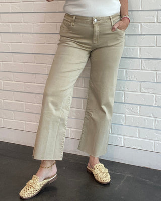 Woman wearing a white tank top and beige pants against a light brick wall.