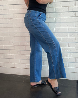 A woman wearing blue wide-leg jeans, a black sleeveless top, and black flat shoes, posing against a white brick wall.