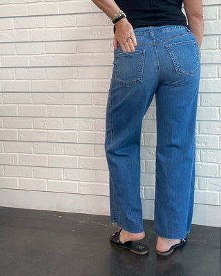 A woman wearing blue straight-leg jeans and a black sleeveless top, shown from the side against a white brick-patterned wall.