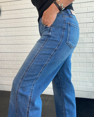 A close-up of a person wearing medium-wash blue denim jeans, showing the back pocket and side seam details, with a black shirt and bracelets visible.