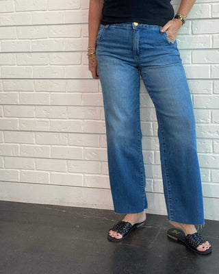A woman wearing a black sleeveless top and blue wide-leg jeans, standing against a white brick wall.
