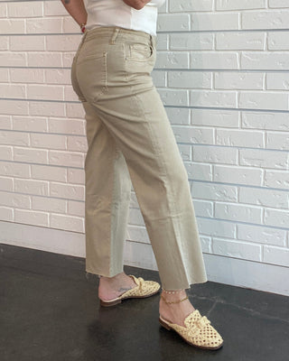 Woman in a white sleeveless top and beige pants standing against a white brick wall.