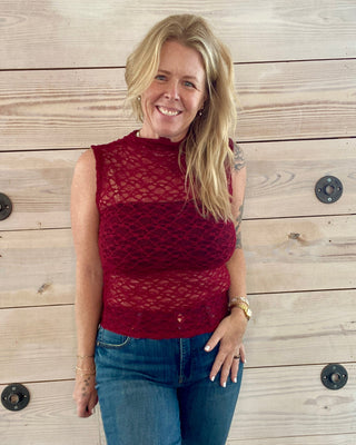 Woman wearing a red sleeveless top and blue jeans standing against a wooden wall.