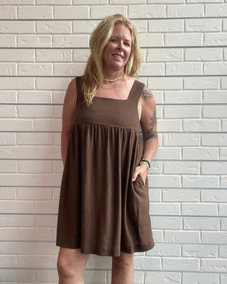 Woman wearing a brown dress standing against a white brick wall.