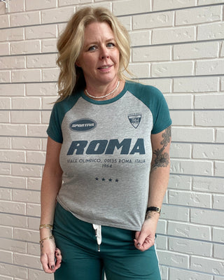 Woman wearing a gray and green 'Roma' t-shirt standing against a light brick wall.