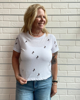 Woman wearing a white t-shirt with small patterns against a light brick wall.