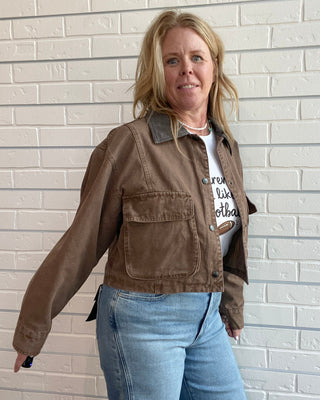 A woman wearing a brown cropped utility jacket with a dark collar, a white graphic t-shirt, and light blue jeans, standing against a white brick wall.