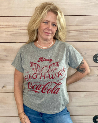 Woman wearing a gray t-shirt with red text standing against a wooden wall.