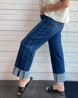 A woman modeling a pair of wide-leg cropped blue jeans with rolled cuffs, paired with a cream sleeveless top and black slide sandals, standing against a white brick wall.