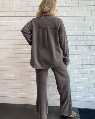 A woman stands with her back to the camera wearing a matching dark brown lounge set consisting of a long-sleeve button-up shirt and wide-leg pants, set against a white brick wall.