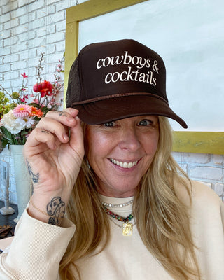 Person wearing a brown cap with 'cowboys & cocktails' text, smiling indoors.