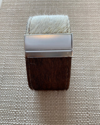A minimalist rectangular ring featuring a polished silver-toned metal band, with one end wrapped in dark brown hair and the other in white hair, displayed on a textured beige woven surface.