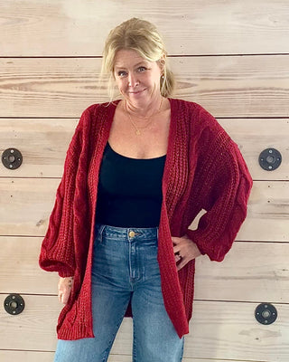 Woman wearing a red cardigan, black top, and blue jeans standing against a wooden wall.