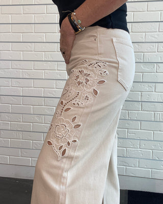 A person wearing off-white eyelet lace cropped pants with floral embroidery on the leg, paired with a black top, shown against a white brick wall background.