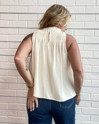 A woman viewed from behind wearing a sleeveless cream-colored top with a smocked neckline and blue jeans, standing against a white brick wall.