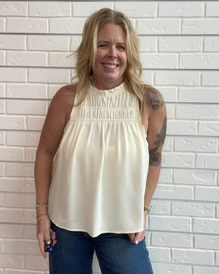 A smiling blonde woman wearing a cream sleeveless halter top and blue jeans, standing against a white painted brick wall.