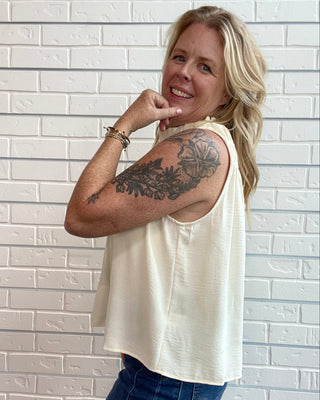 A woman with blonde hair and a floral arm tattoo models a cream-colored sleeveless top and blue jeans, posing in front of a white brick wall.