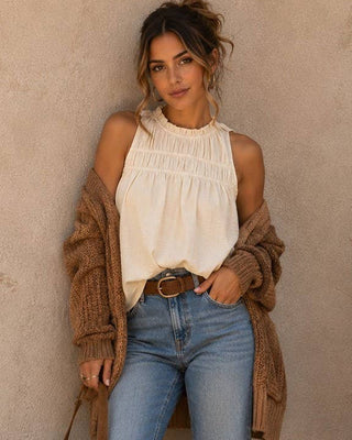 A woman posing in a cream sleeveless smocked blouse, a brown knit cardigan, blue jeans, and a brown belt, standing against a textured beige stucco wall.