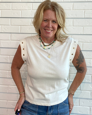 A smiling blonde woman wearing a sleeveless cream-colored top with gold button details on the shoulders and blue jeans, standing against a white brick wall.