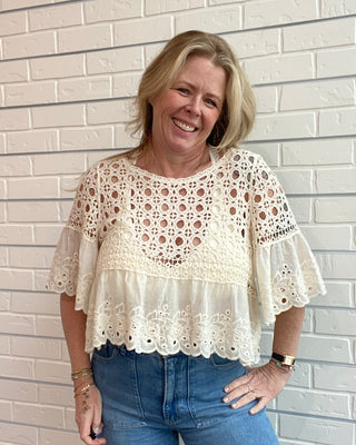 Woman wearing a white lace blouse and blue jeans standing against a light brick wall.