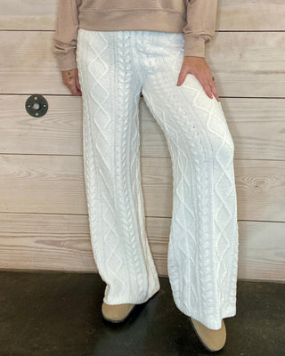 Person wearing a beige sweatshirt with text and white pants against a wooden background