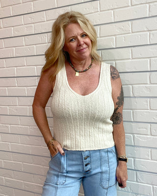 Woman wearing a white tank top and blue jeans standing against a light brick wall.