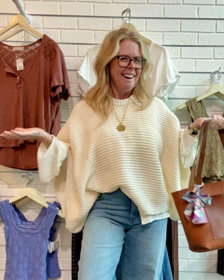Woman in a clothing store wearing a beige sweater and blue jeans, holding a brown handbag.