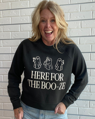 A woman with blonde hair smiling in front of a white brick wall, wearing a black crewneck sweatshirt with white text reading 'HERE FOR THE BOO-ZE' and three ghost illustrations, paired with light blue denim shorts.