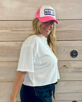 Woman wearing a white shirt, black shorts, and a pink and white cap against a wooden wall.