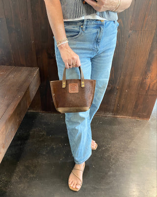 A person wearing light blue jeans and beige sandals holds a brown leather handbag with gold-tone hardware in front of a dark wood-paneled wall.