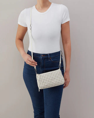 Person wearing a white shirt and blue jeans holding a woven handbag against a plain background