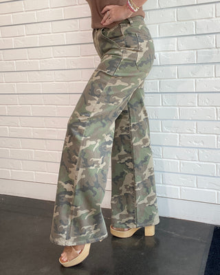 Person wearing camouflage pants and a brown top against a white brick wall.