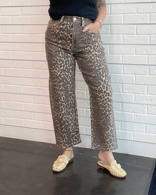 Woman wearing a black tank top and leopard print pants against a white brick wall.