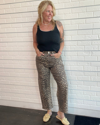 Woman wearing a black tank top and leopard print pants standing against a white brick wall.