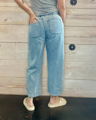 Person wearing a gray t-shirt and blue jeans standing against a wooden wall.