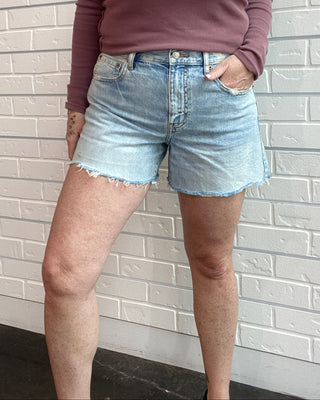 Person wearing a mauve long-sleeve top and light blue denim shorts against a white brick wall.