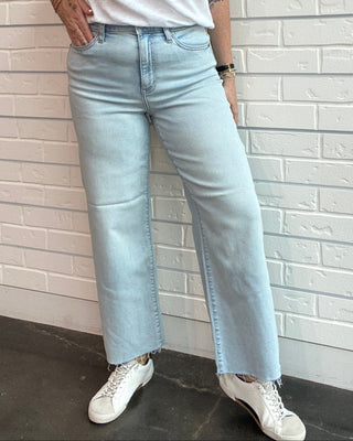 Person wearing a white t-shirt, light blue jeans, and white sneakers against a white brick wall.