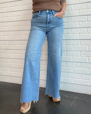 Woman wearing a brown tank top and blue jeans standing against a white brick wall.