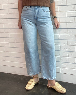 Woman wearing a brown tank top and light blue jeans standing against a white brick wall.