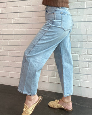 Woman posing against a white brick wall wearing a brown sleeveless top and light blue jeans.