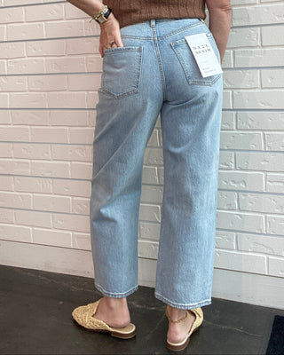 Person wearing a brown sleeveless top and light blue jeans with a tag, standing against a light brick wall.