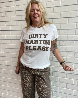 A smiling blonde woman stands against a white brick wall wearing a white graphic t-shirt with the text 'DIRTY MARTINI PLEASE' in brown block letters, paired with leopard-print pants.