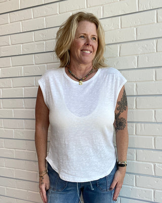 Woman wearing a white sleeveless top and blue jeans standing against a light brick wall.