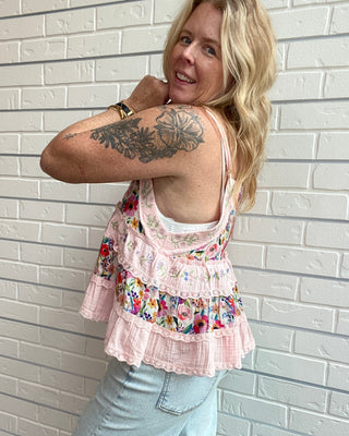A woman wearing a light pink tiered ruffle tank top with a colorful floral print along the underbust band, paired with light blue jeans, posing against a white brick wall.